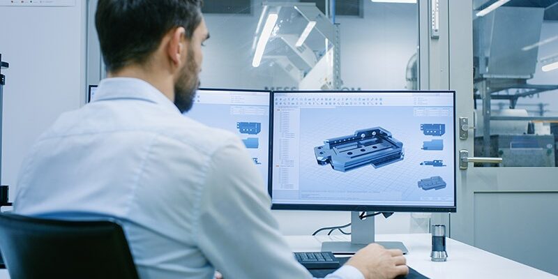 Engineer/ Technician Working on a Personal Computer with Two Displays, He's Designing New Component in CAD Program. Out of the Office Window Components Manufacturing Factory is Seen.